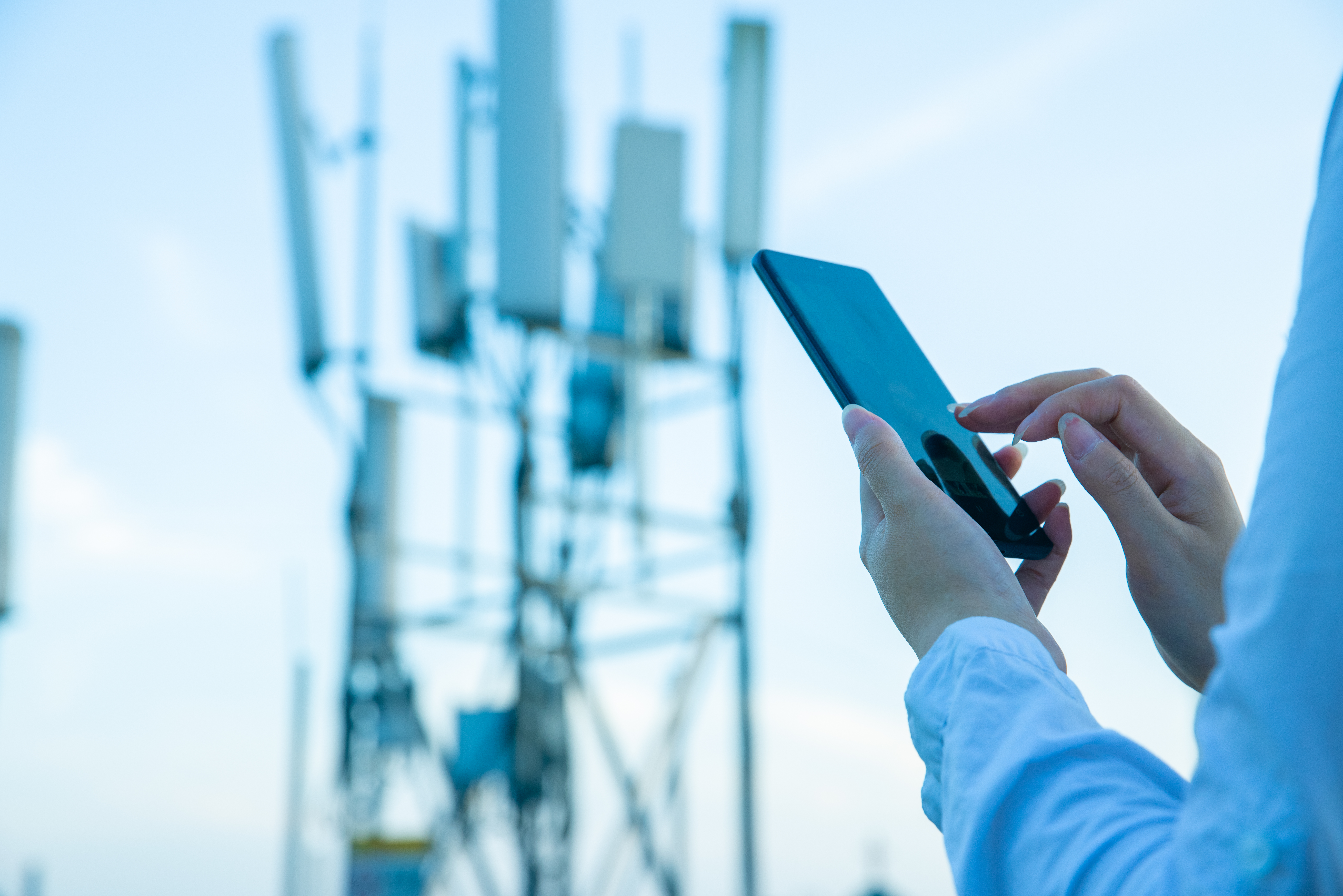A person uses a smartphone outdoors with cellular antennas and communication towers in the background against a clear sky.