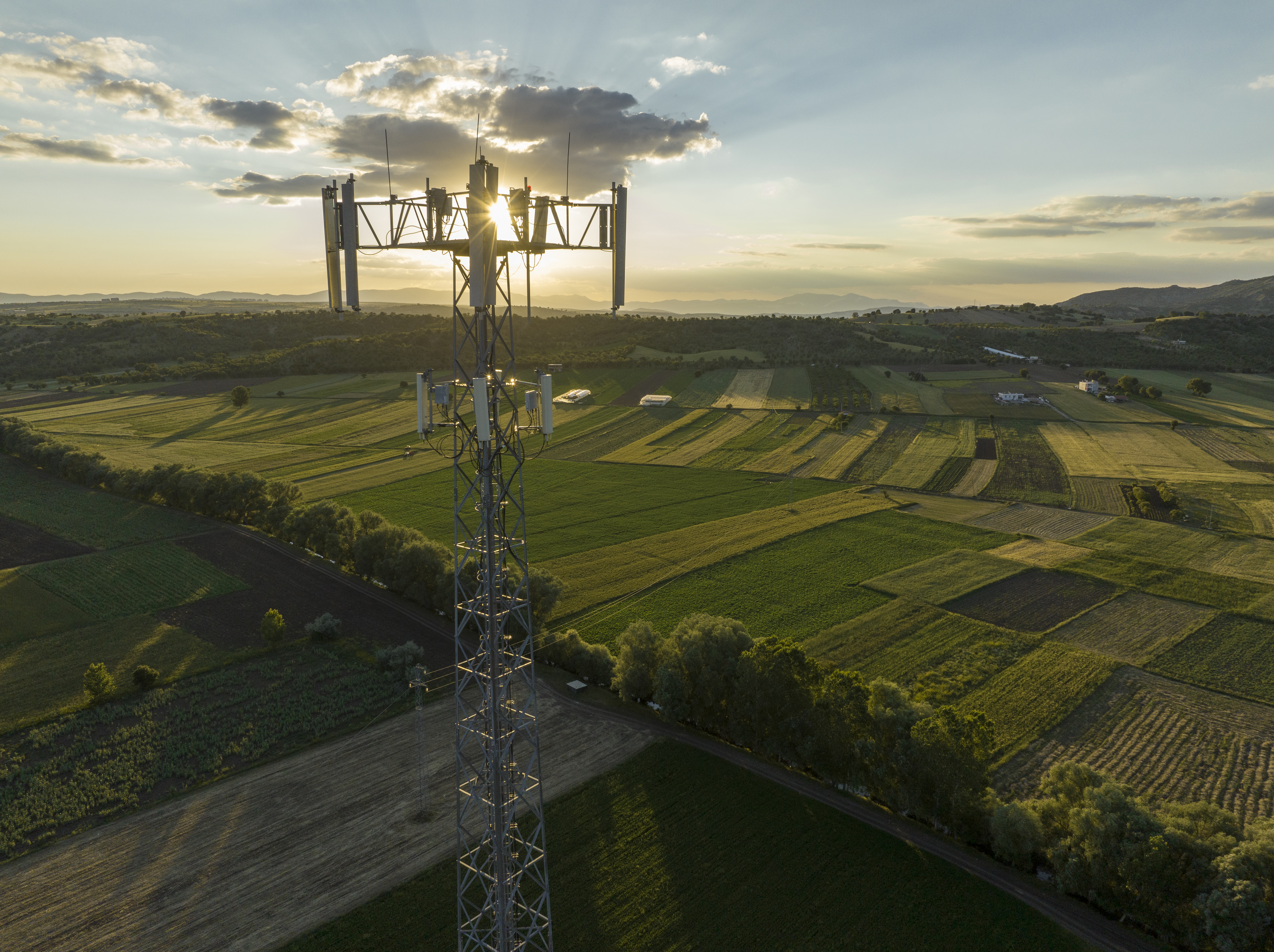 An aerial view of a large cellular telecommunications tower rising above lush green farmland and fields at sunset.