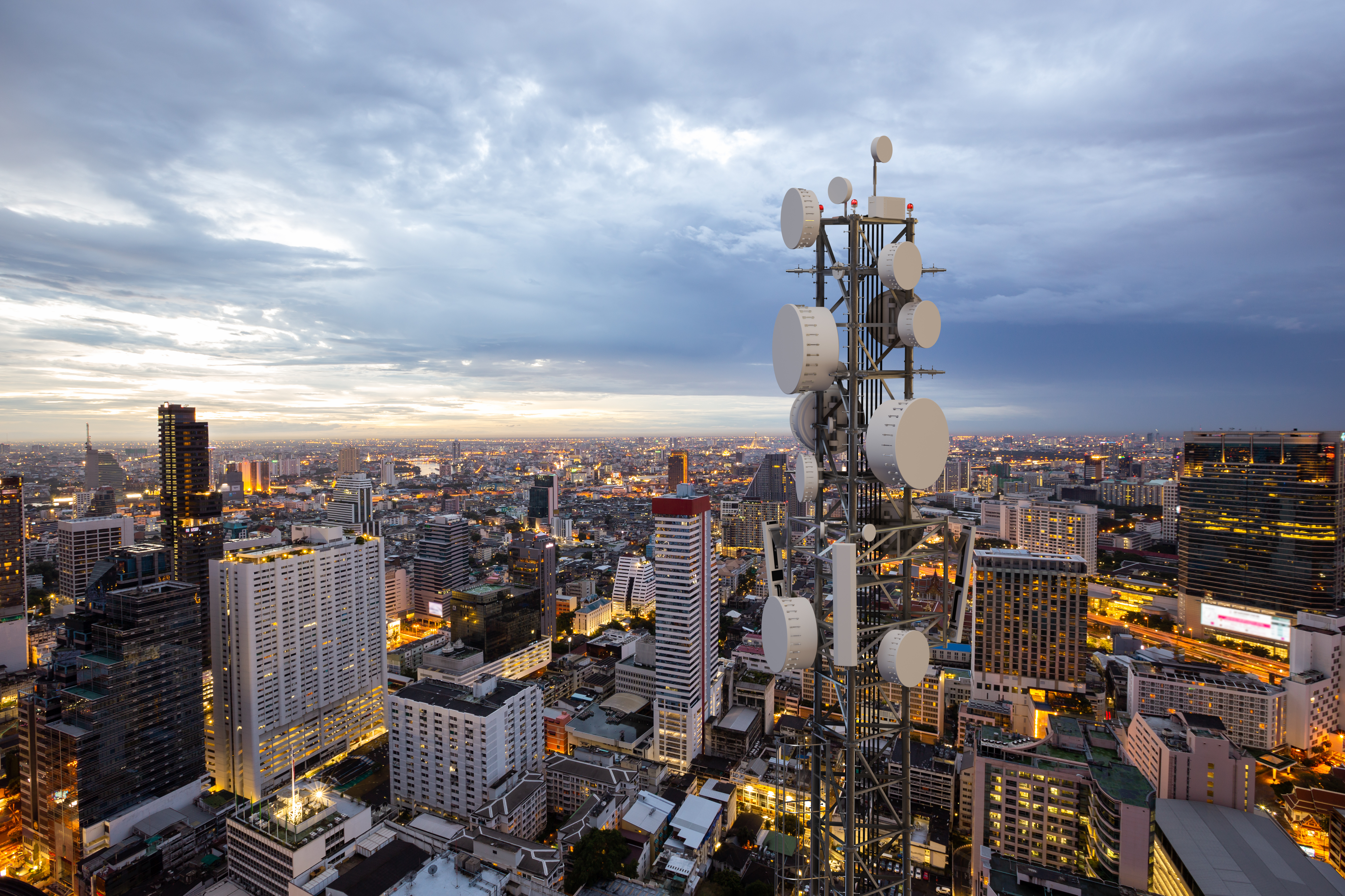 An elevated view of a large communication tower with multiple white satellite dishes overlooking a dense city skyline at dusk.