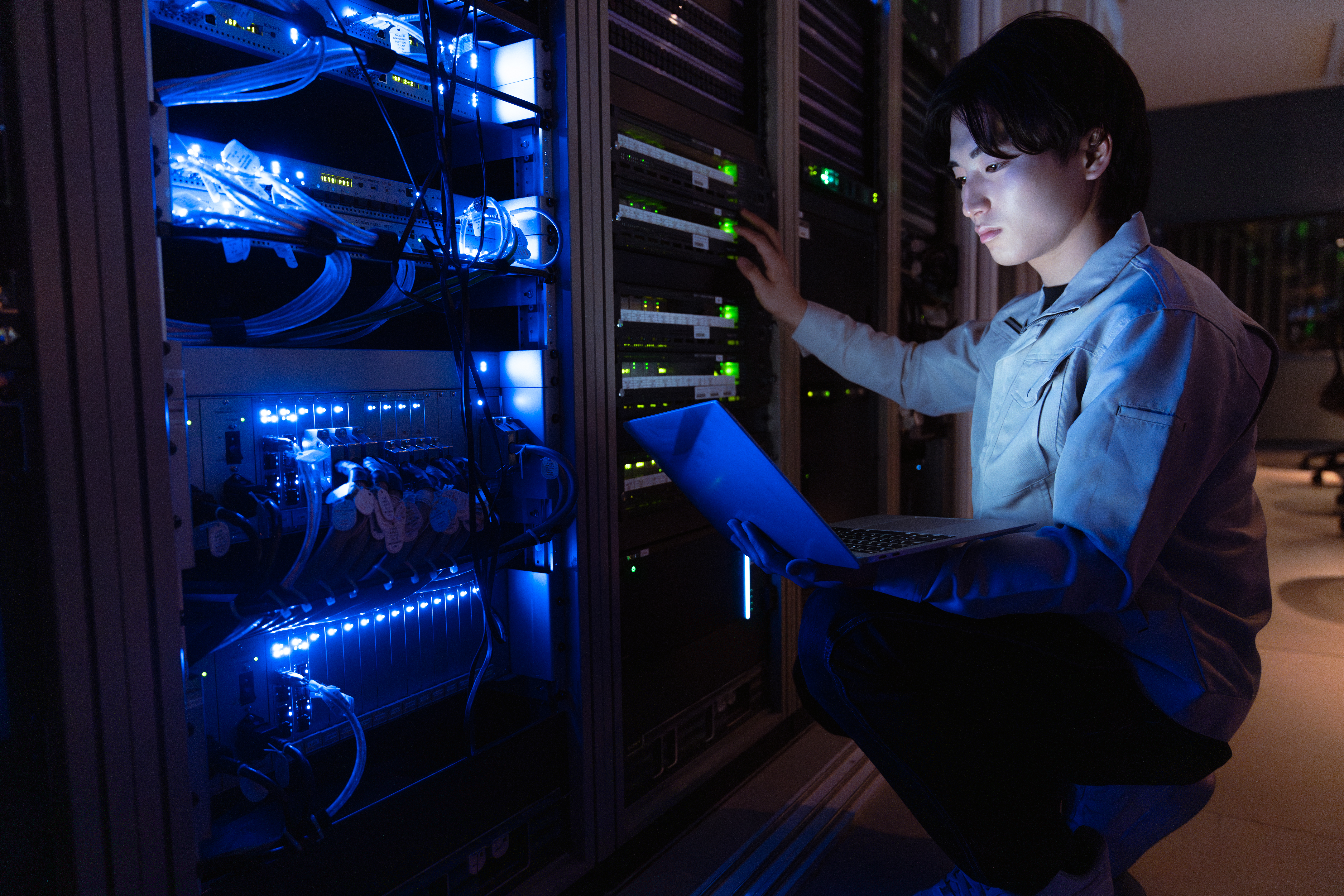 Focused technician kneeling in a dark server room, working on a laptop while checking glowing blue network racks.