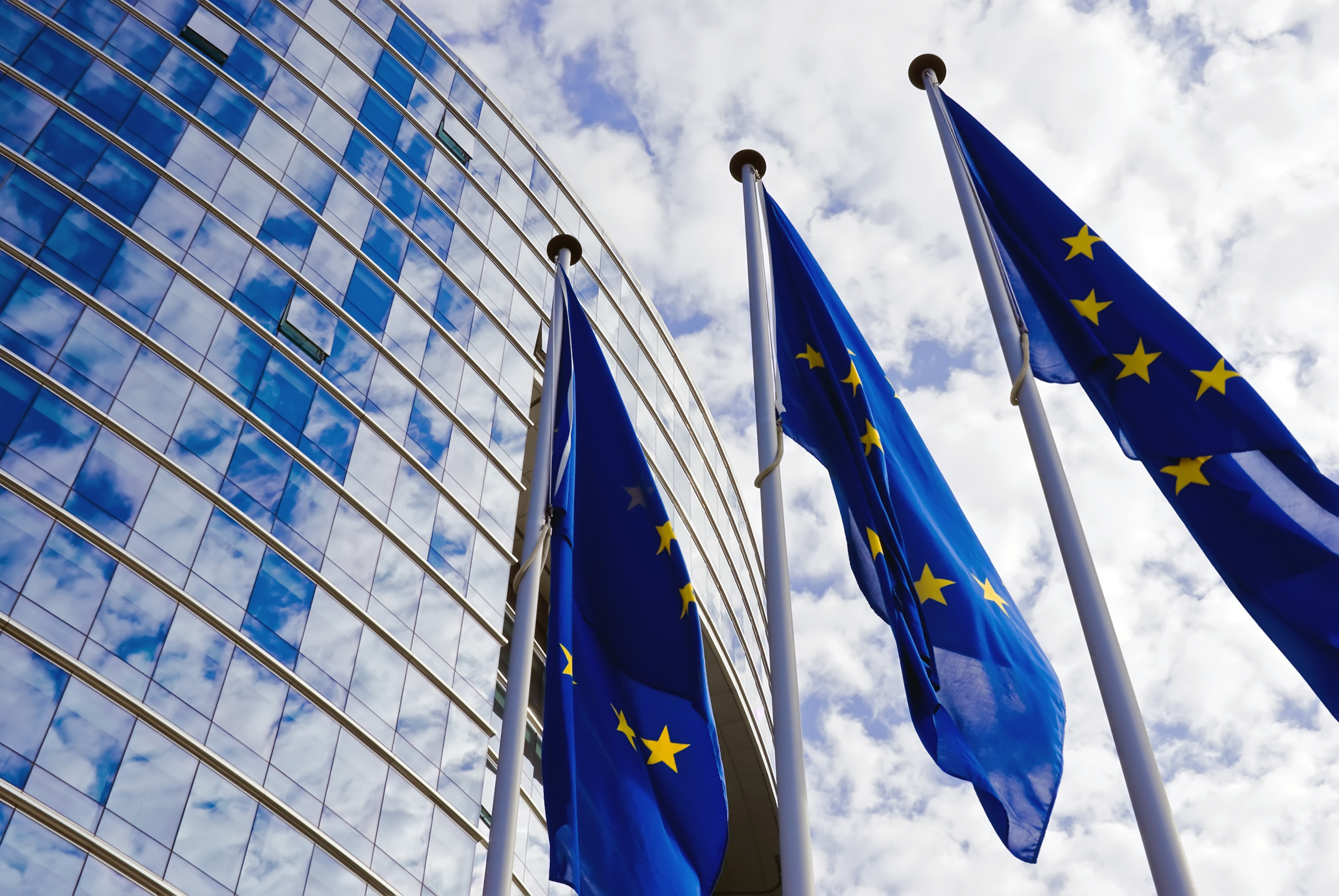 Three EU flags wave in front of a modern glass building under a partly cloudy sky.