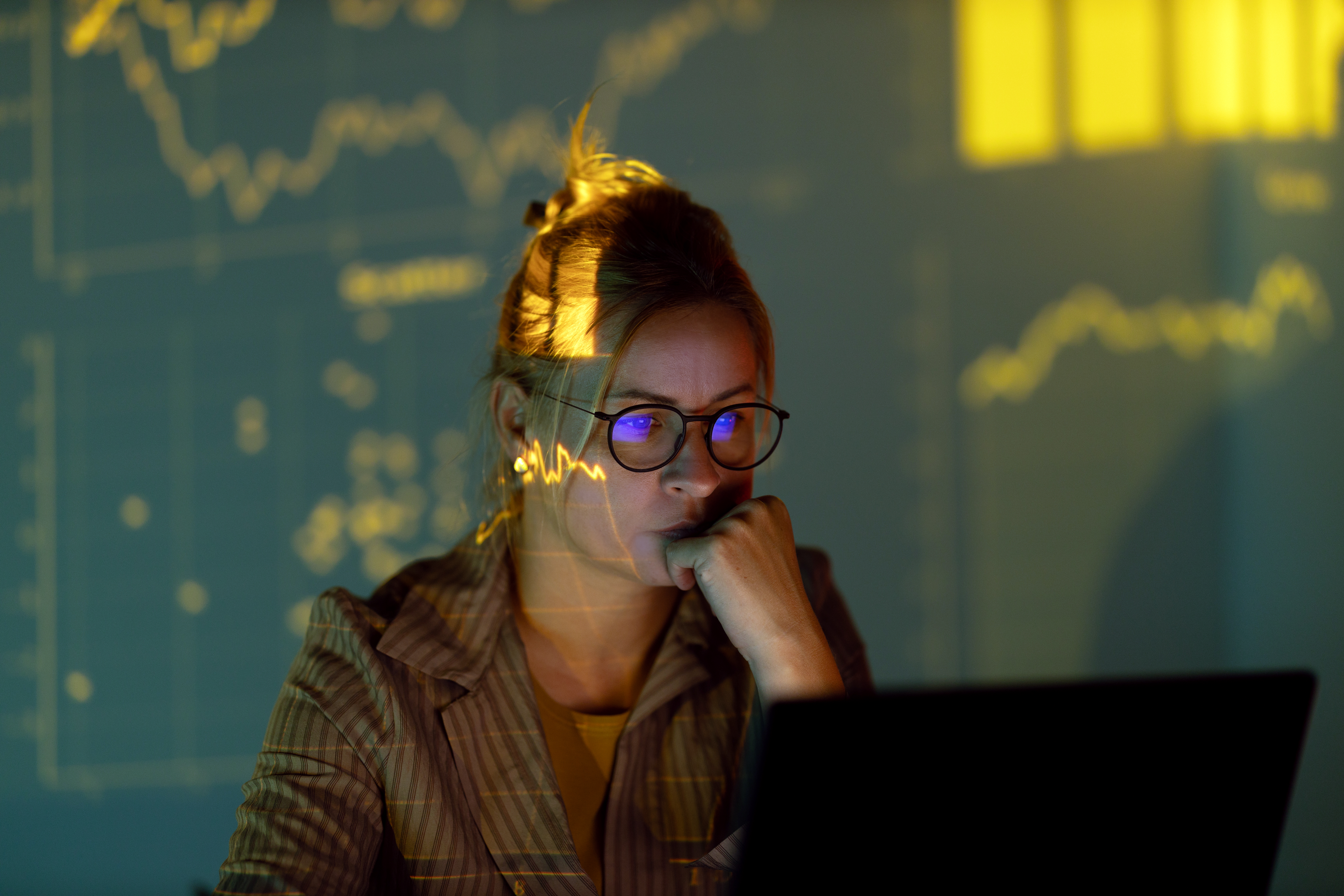 A focused data analyst wearing glasses works on a laptop in a dimly lit room, with glowing yellow stock charts or graphs projected onto her face and the wall.
