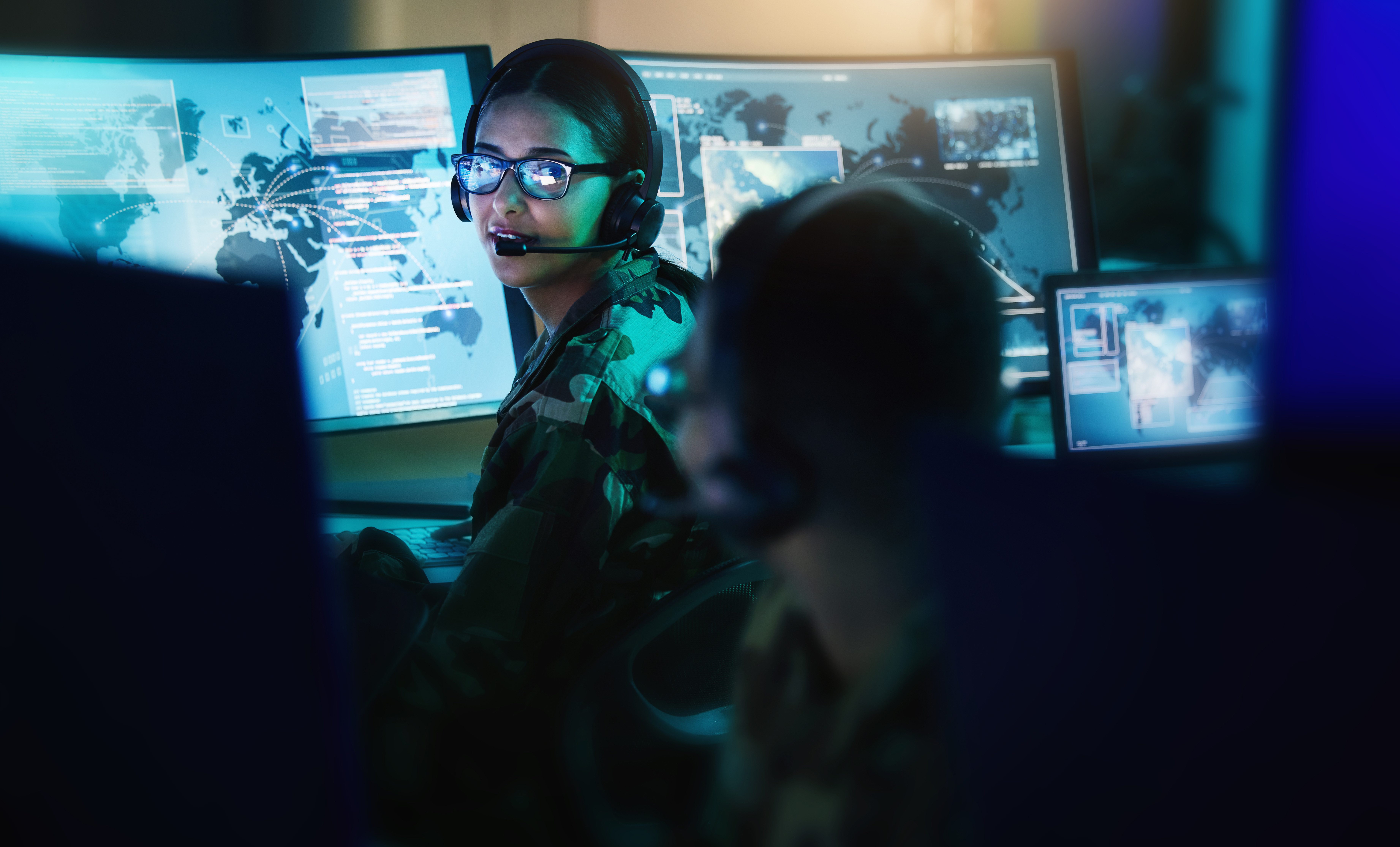 A man and a woman in military uniforms, wearing headsets, work in a dimly lit control room, monitoring digital maps and data on multiple screens.