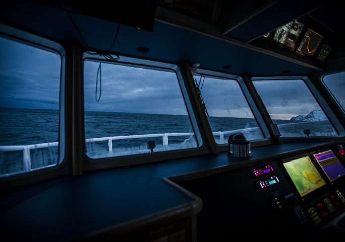 View from inside a ship’s bridge showing navigation screens and the ocean through wide front windows, at dusk.