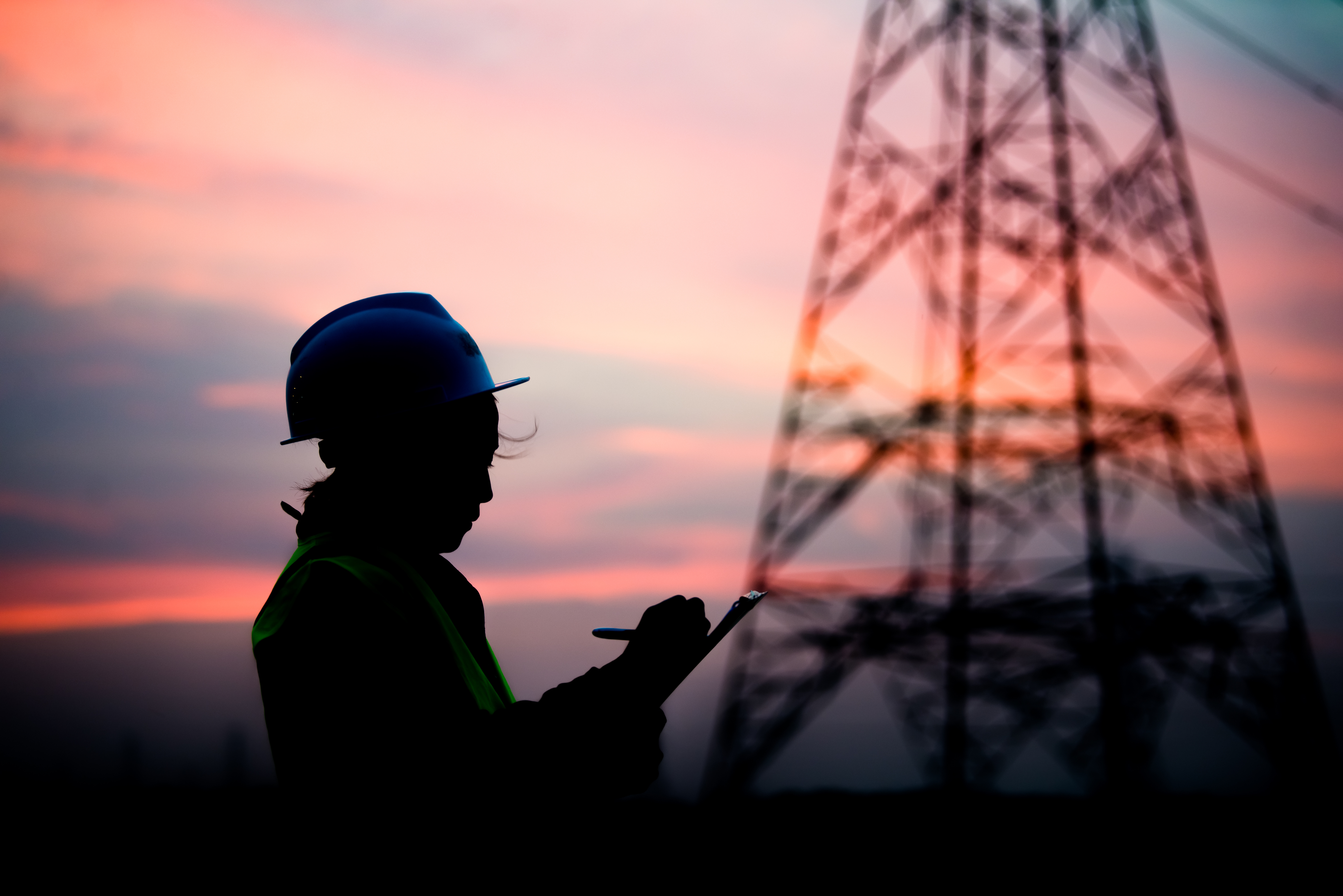 Silhouette of an engineer wearing a safety helmet and reflective vest, writing on a clipboard near a power transmission tower at sunset.