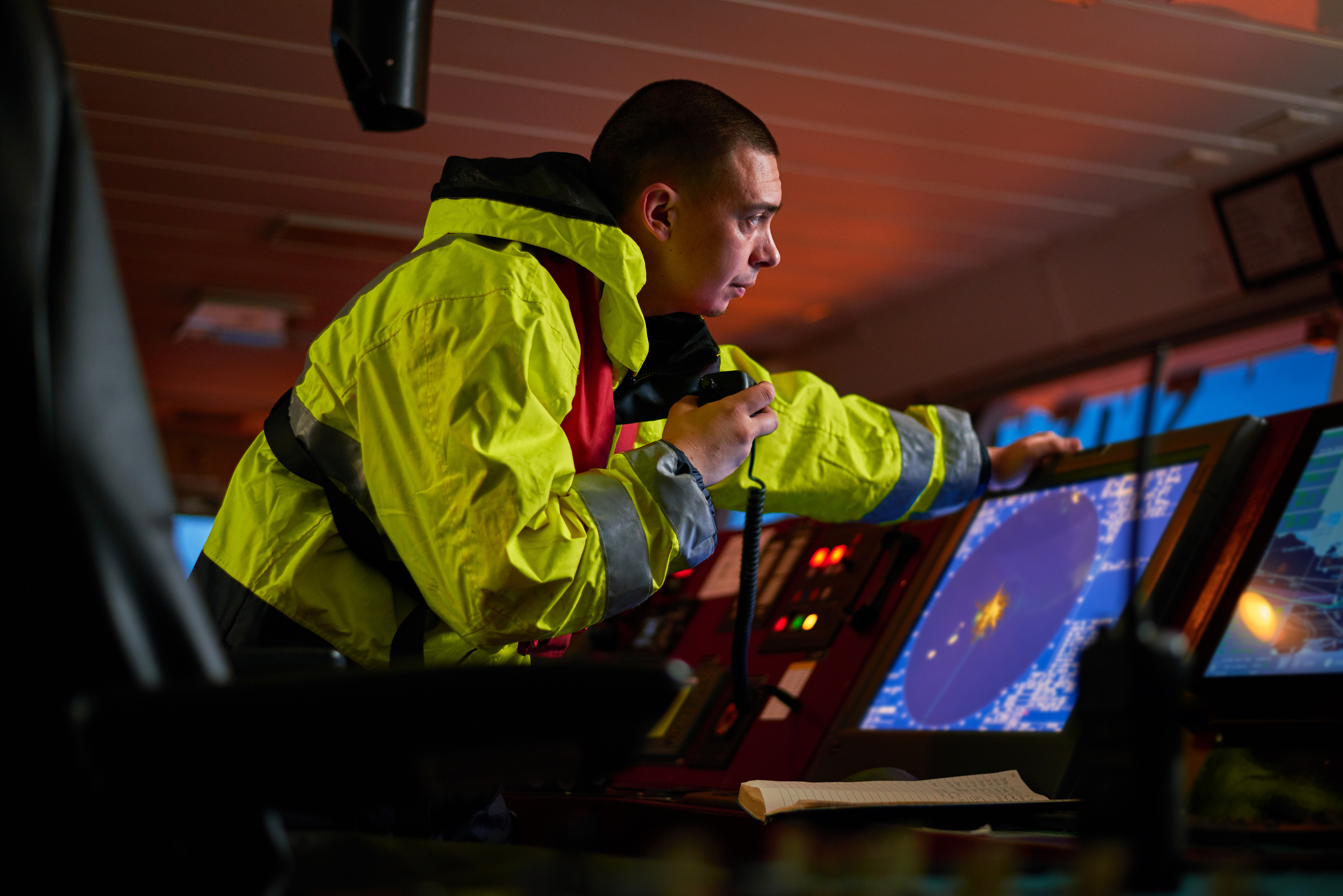 A crew member in a yellow jacket operates navigation equipment on a ship’s bridge, monitoring digital radar screens.