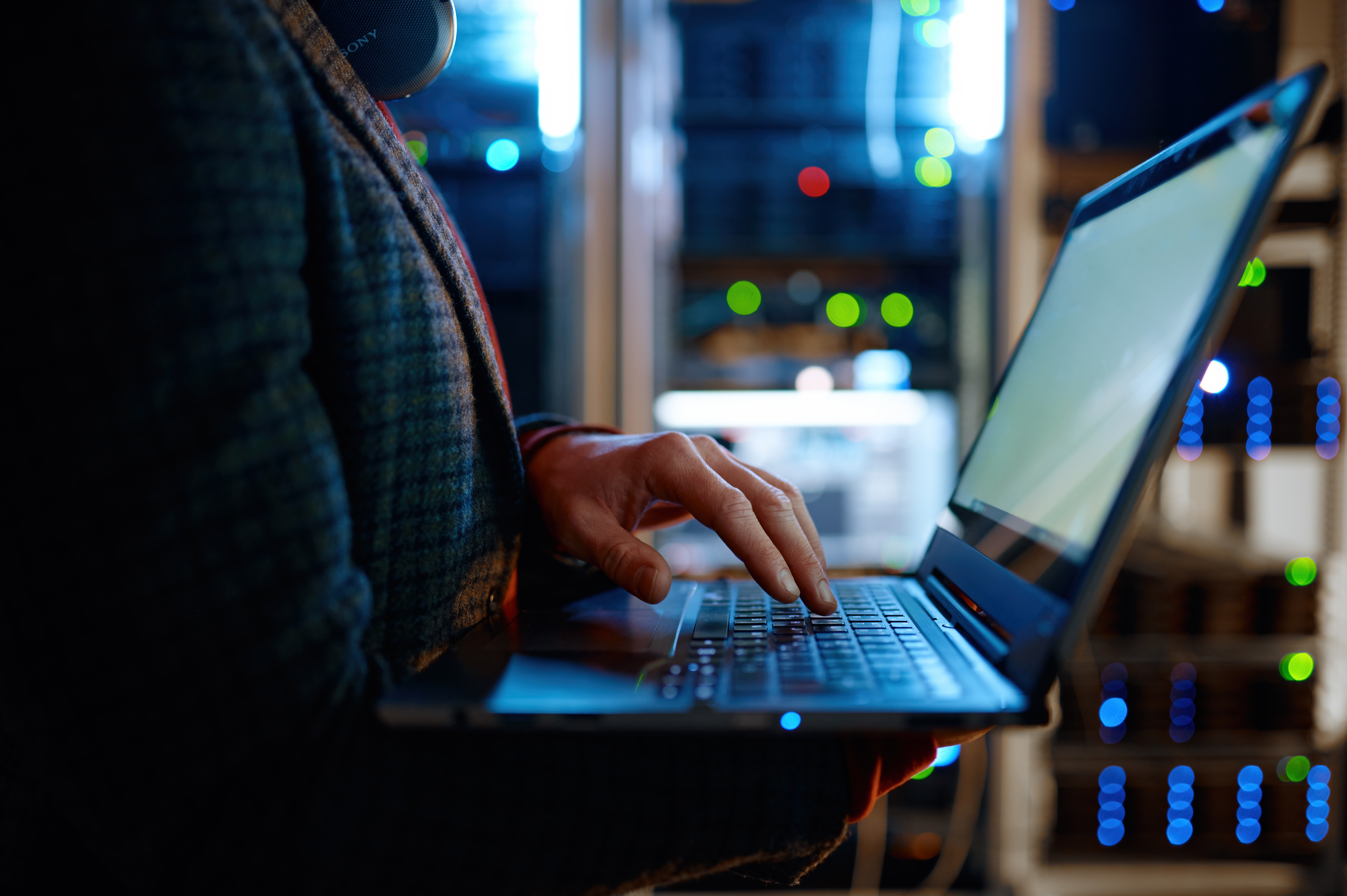 A person types on a laptop in a server room with glowing blue and green lights from network equipment.
