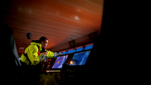 A crew member in a yellow jacket operates navigation equipment on a ship’s bridge, monitoring digital radar screens.