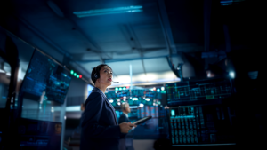 A woman wearing a headset and holding a tablet looks up while monitoring multiple screens in a dimly lit control room